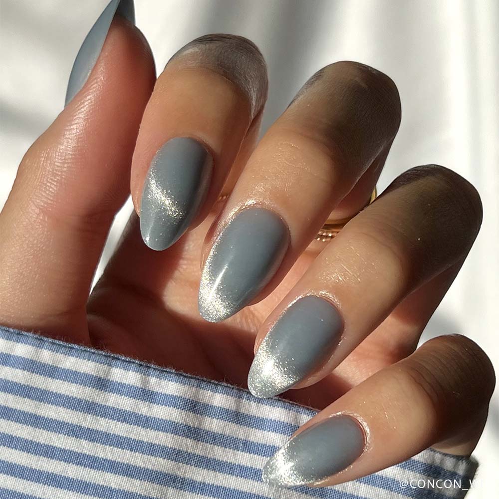 A hand with almond-shaped nails painted in glamnetics Baby Blues—matte gray with shiny silver tips and a velvet metallic finish—rests against a blue and white striped shirt, soft sunlight highlighting the look.