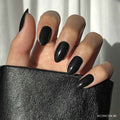 A hand with long, glossy black glamnetic Boba vegan cruelty-free nails holds a textured black leather item in bright lighting, with a white curtain in the background.