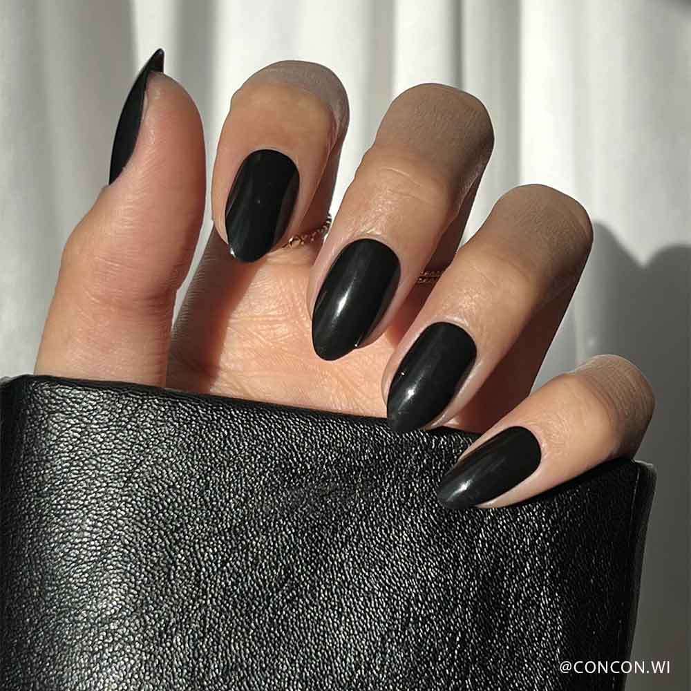 A hand with long, glossy black glamnetic Boba vegan cruelty-free nails holds a textured black leather item in bright lighting, with a white curtain in the background.