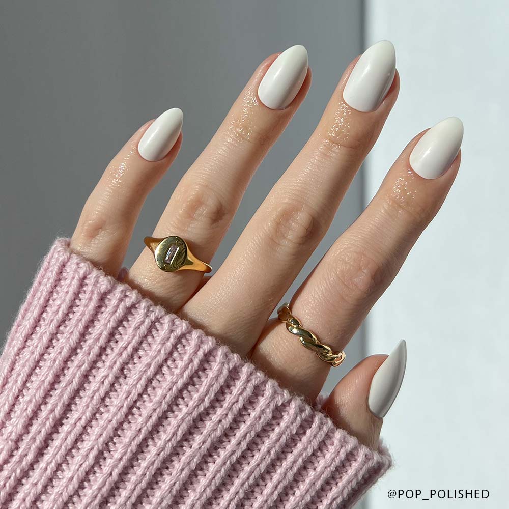 A hand with almond-shaped, glossy white Angel vegan press-on nails by glamnetic, adorned with two gold rings, is shown against a neutral background. The person wears a pink ribbed sweater with the sleeve partially covering their fingers.