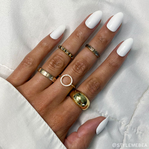 A hand wears glamnetic Angel long, white, almond-shaped press-on nails and five textured gold rings. The person is dressed in a white long-sleeved shirt against a soft white fabric background.