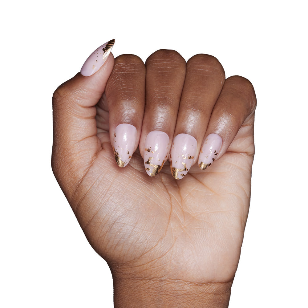 A hand with medium brown skin shows off short, almond-shaped nails in glamnetic’s 24K set—pale pink polish with gold foil-tipped accents—set against a white background.