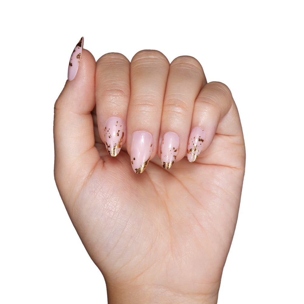 A hand with short almond-shaped nails painted in glamnetics 24K, a light pink shade with gold foil tip accents, is shown against a white background.
