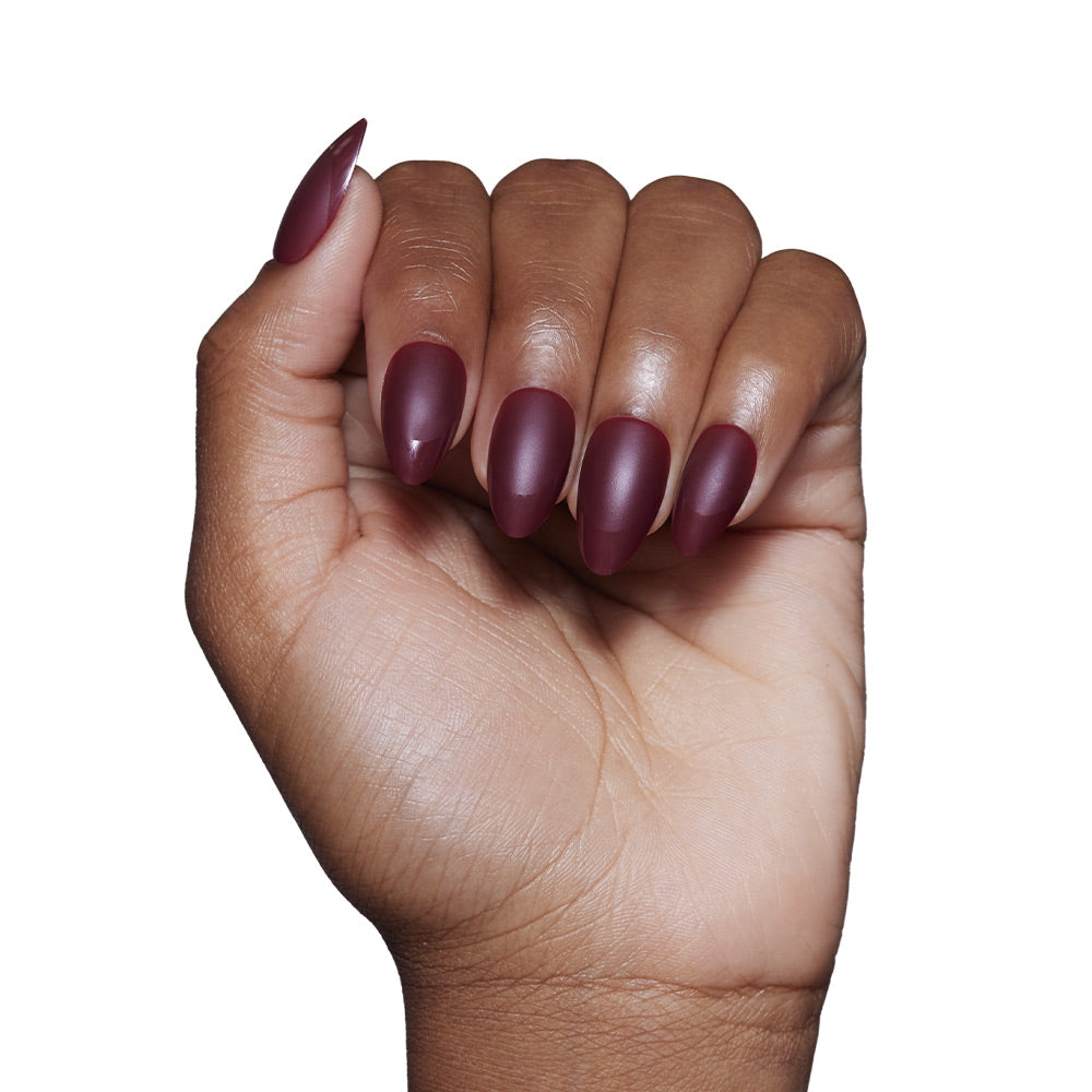 A hand with medium brown skin displays long, almond-shaped nails in the glamnetic Merlot matte set. The fingers are slightly curled against a plain white background.
