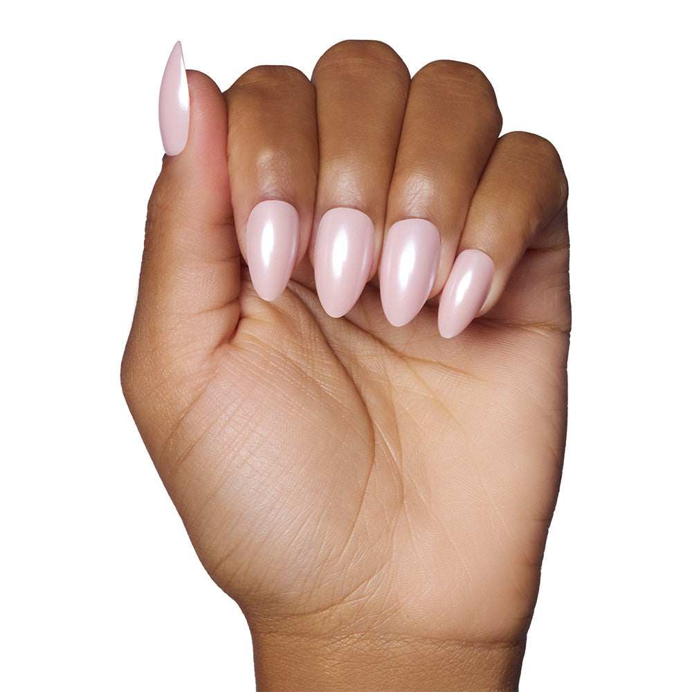 A hand with medium-brown skin shows short almond-shaped nails painted in a glossy blush pink using glamnetic’s First Kiss, set against a white background.