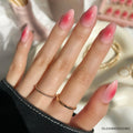 A hand with a short almond shape showcases a pink ombré design inspired by the Blushing Aura nail set from glamnetic, paired with two delicate gold rings and a dark sweater. Jewelry and nail polish bottles are blurred in the background.