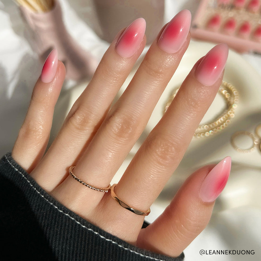 A hand with a short almond shape showcases a pink ombré design inspired by the Blushing Aura nail set from glamnetic, paired with two delicate gold rings and a dark sweater. Jewelry and nail polish bottles are blurred in the background.