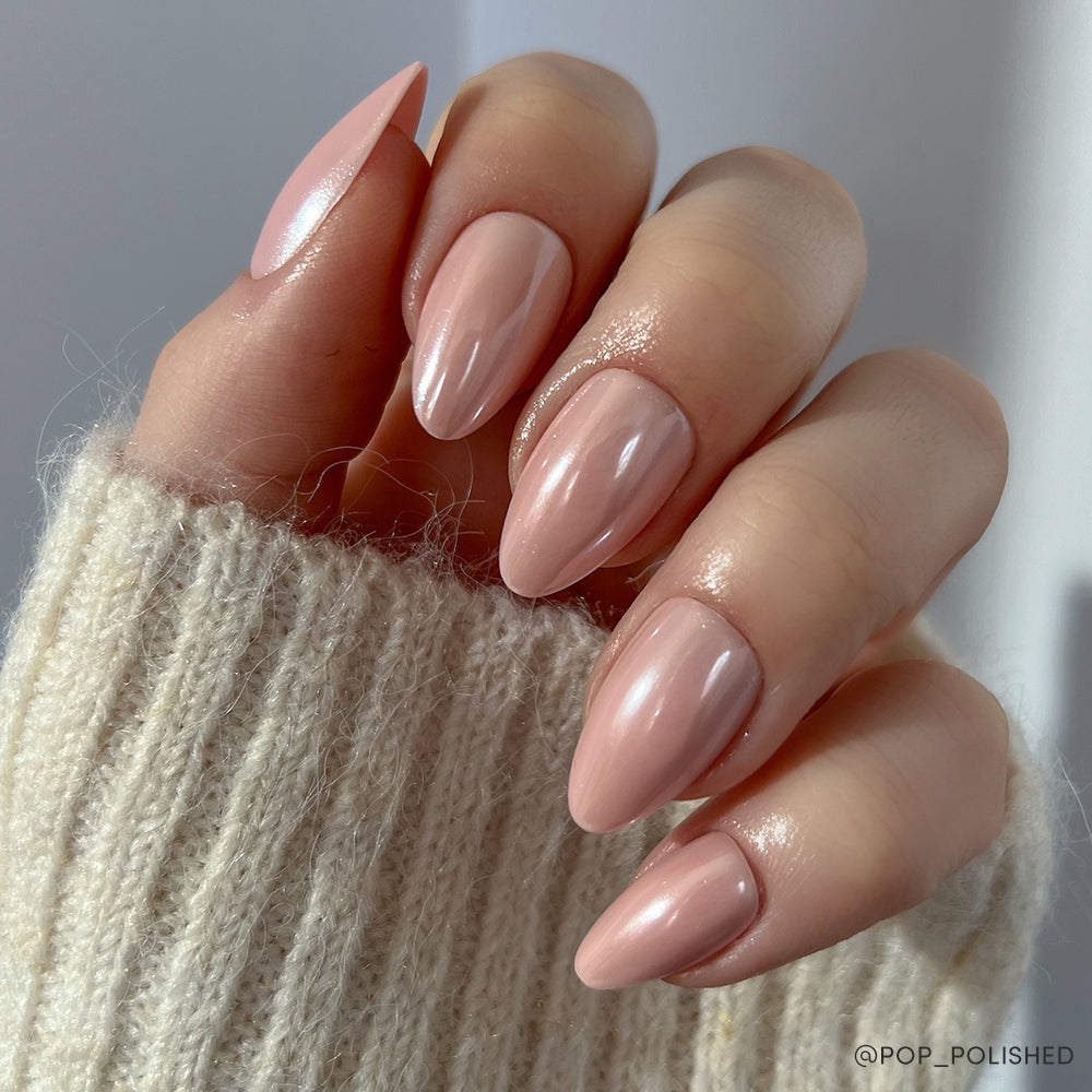 A hand with short almond-shaped nails in a glossy blush pink, possibly using the glamnetic First Kiss set, is shown against a soft light background. The person is wearing a cream-colored knit sweater.