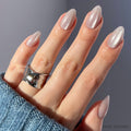 A hand showcases the glamnetic Selenite Nail Set in a short almond shape with shimmery sheer pink polish. A chunky silver ring and blue knit sweater complete the look, while soft lighting highlights the velvet finish of the nails.