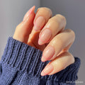 A hand wears glamnetic’s Hot Take short almond nails with a sheer pink base and bright pink French tips, styled with a blue knit sweater.