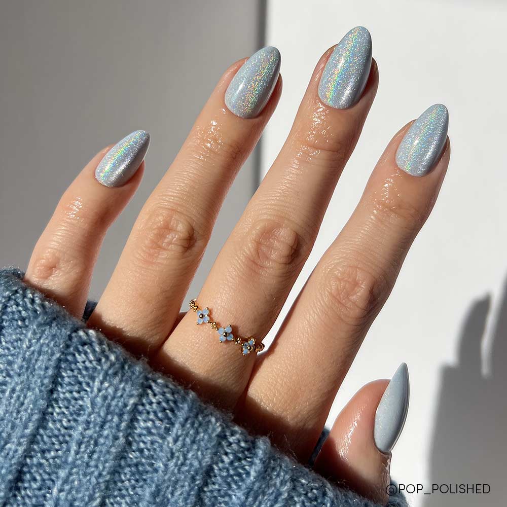 The Frozen nail set by glamnetic features short almond-shaped nails in an icy-blue shade. The hand is accessorized with a delicate gold ring with blue stones and partially covered by a blue knit sweater.
