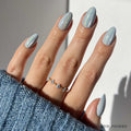 The Frozen nail set by glamnetic features short almond-shaped nails in an icy-blue shade. The hand is accessorized with a delicate gold ring with blue stones and partially covered by a blue knit sweater.