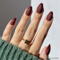Wearing the Merlot press-on nails by glamnetic, this hand features short almond-shaped nails with a matte finish and glossy burgundy tips, styled with a gold green gemstone ring and a green knit sweater.