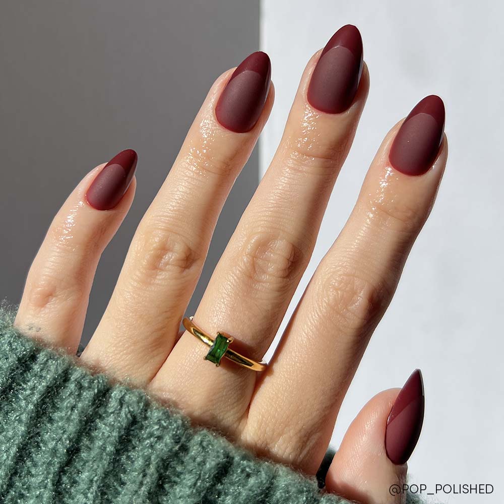 Wearing the Merlot press-on nails by glamnetic, this hand features short almond-shaped nails with a matte finish and glossy burgundy tips, styled with a gold green gemstone ring and a green knit sweater.