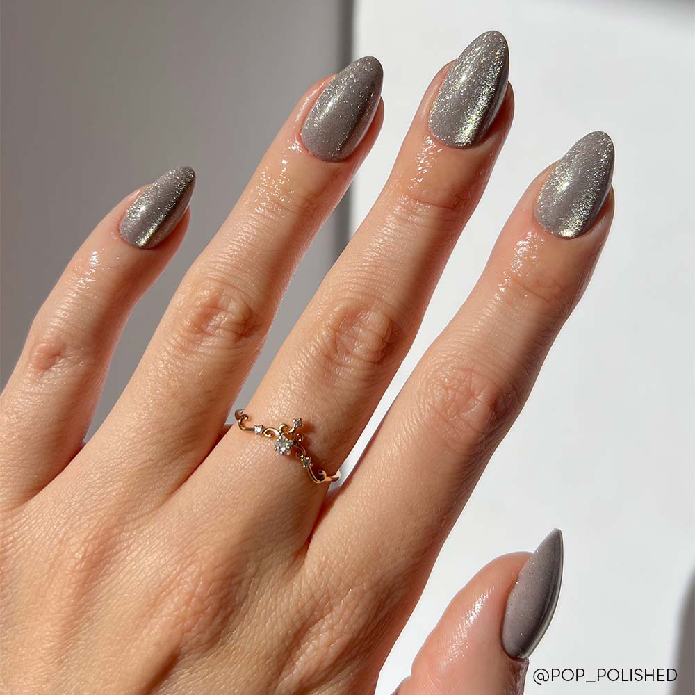 A hand with short-almond nails painted in glamnetic’s Oslo, a shimmery silver cat-eye, wears a delicate gold ring with small gemstones. The fingers are slightly spread against a plain, light background.