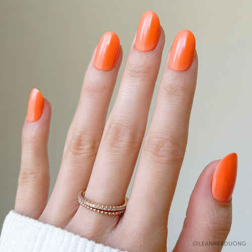 A hand with long, almond-shaped Apricot press-on nails by glamnetic is shown in vibrant orange. Two gold rings accent the ring finger, a white sleeve covers the wrist, and the background is softly blurred.