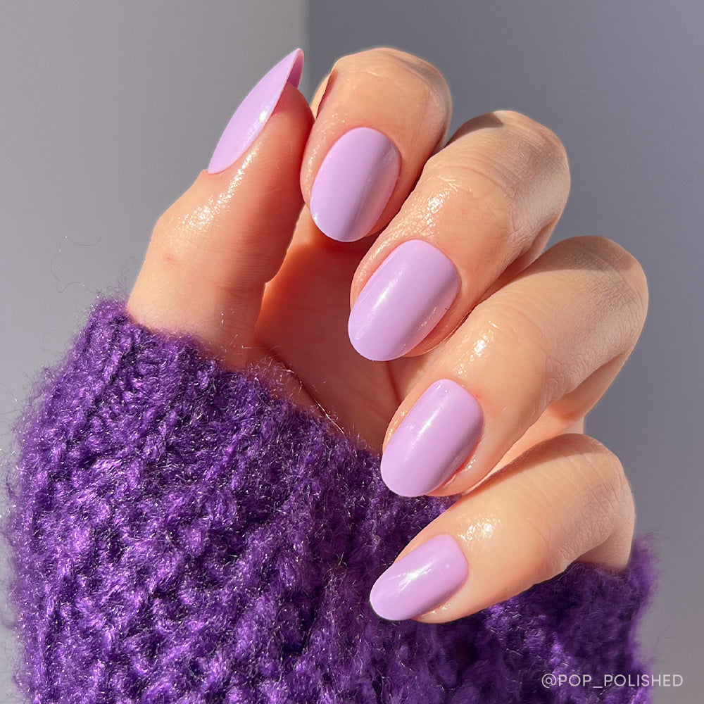 A hand with neatly manicured, oval-shaped nails painted in glamnetic’s Lilac set wears a textured purple knit sweater, shown against a softly lit background.