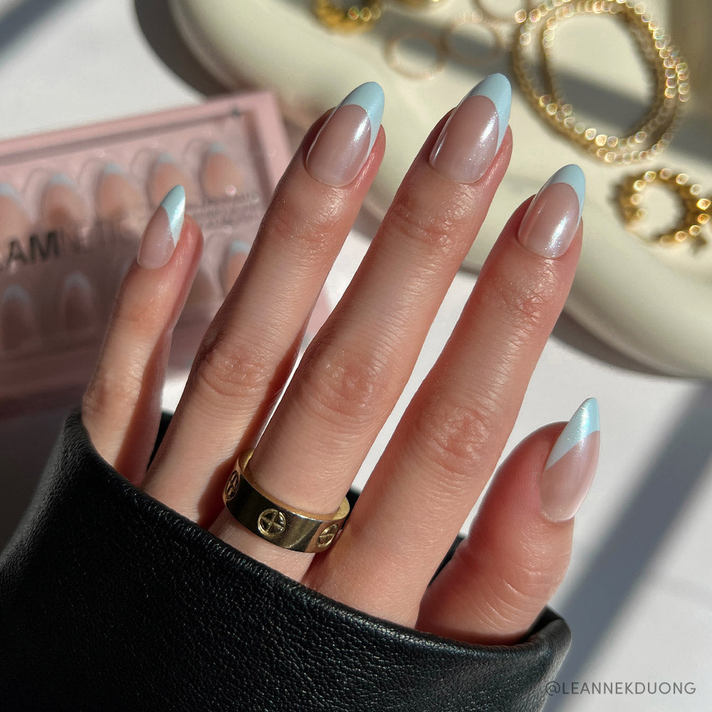 A hand with long, almond-shaped nails in glamnetic’s Blueberry Icing set—baby blue French tips and a glazed finish—wears a gold ring, is partly covered by a black sleeve, with the Blueberry Icing box and gold jewelry in the background.