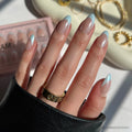 A hand with long, almond-shaped nails in glamnetic’s Blueberry Icing set—baby blue French tips and a glazed finish—wears a gold ring, is partly covered by a black sleeve, with the Blueberry Icing box and gold jewelry in the background.