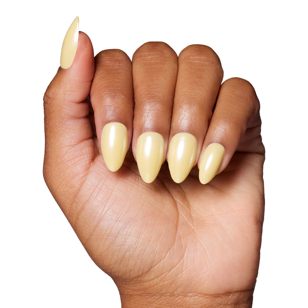A hand with medium brown skin shows long, almond-shaped press-on nails in glossy pastel yellow from the glamnetic Yellow Pop set, displayed against a white background.