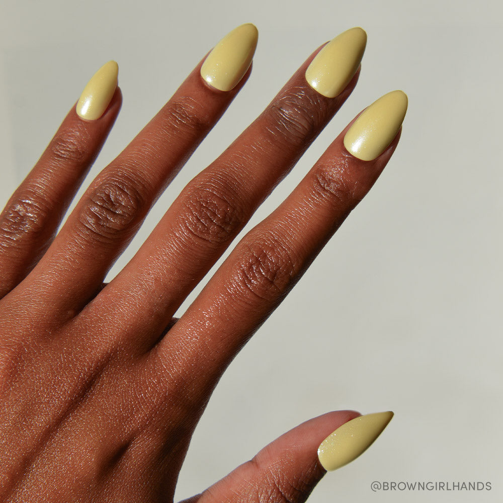 A close-up of a hand with medium-brown skin and long, almond-shaped nails painted in Yellow Pop by glamnetic. The plain, light background highlights the soft charm of this pastel yellow nail set.