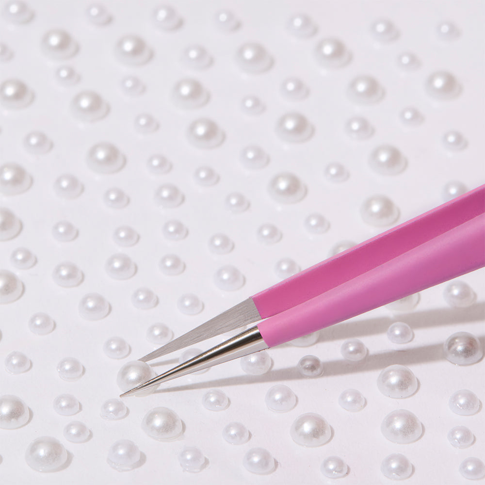 A pair of glamnetic Nail Decal Tweezers with an ultra-precise design picks up a small, round, white pearl from scattered pearls on a white surface.