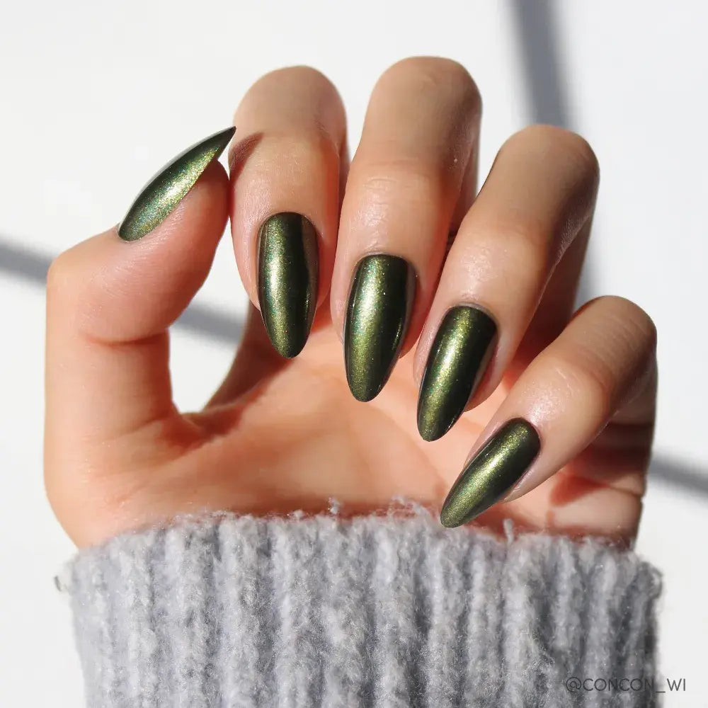 A hand with long, almond nails painted in glamnetic's Cutie Pine—a shimmery dark green polish—rests against a light background. The person is wearing a light gray ribbed sweater.