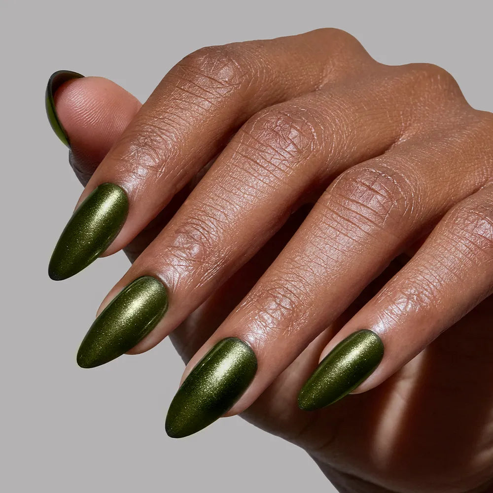 A hand with almond-shaped "Cutie Pine" nails by glamnetic, featuring metallic olive green polish, is displayed against a plain gray background.