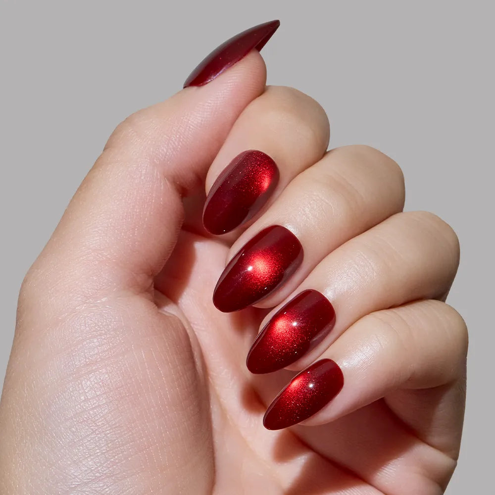A hand with short almond nails, painted in the shiny, metallic red Frosted Cranberry polish by glamnetic, is displayed against a plain gray background.