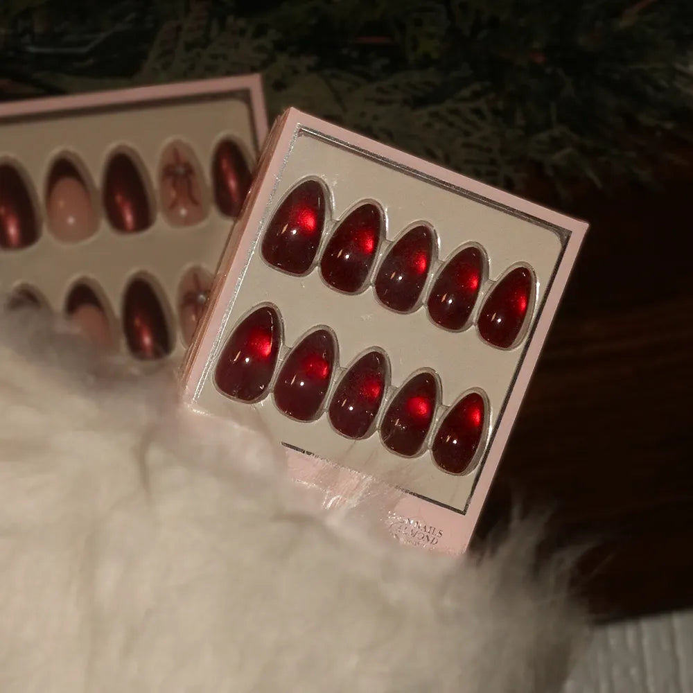 A box of glamnetic Frosted Cranberry glossy red press-on nails is shown partially tucked in a fluffy white stocking, with another festive nail set visible in the background.