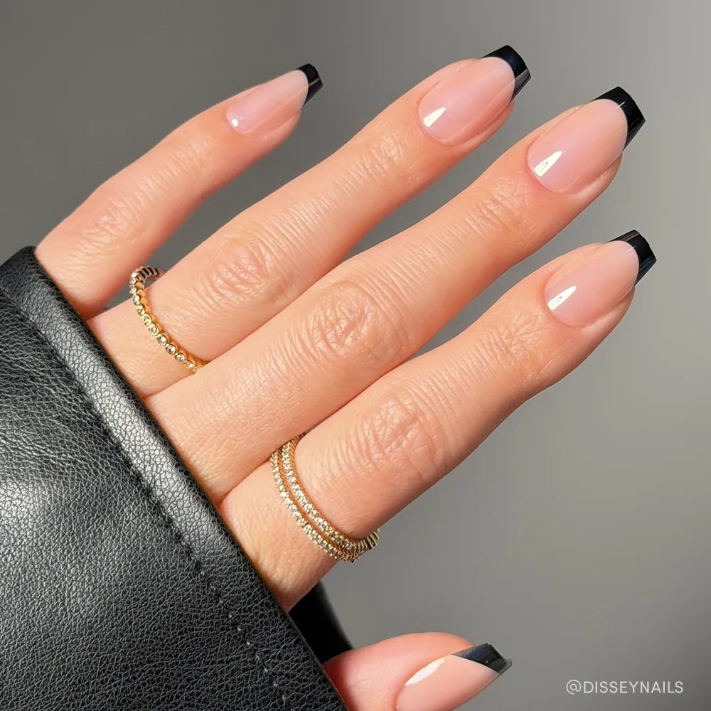 A hand with long, almond-shaped glamnetic Balsamic Glaze press-on nails, showcasing a glossy nude base and sharp black French tips. Two delicate gold rings adorn the fingers, while a black leather sleeve partially covers the hand.