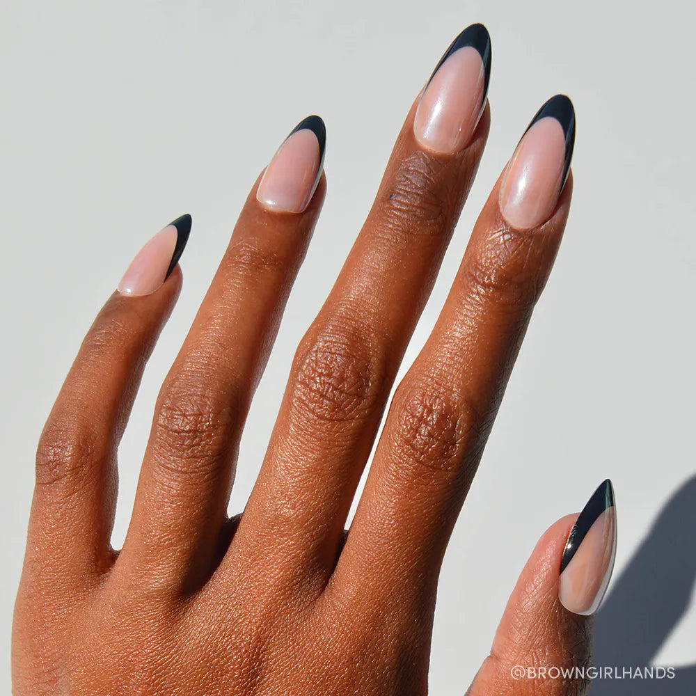 A hand with medium brown skin shows off glamnetic's Black Truffle nails: long, almond-shaped, in a nude shade with sleek black French tips. The background is bright and plain.