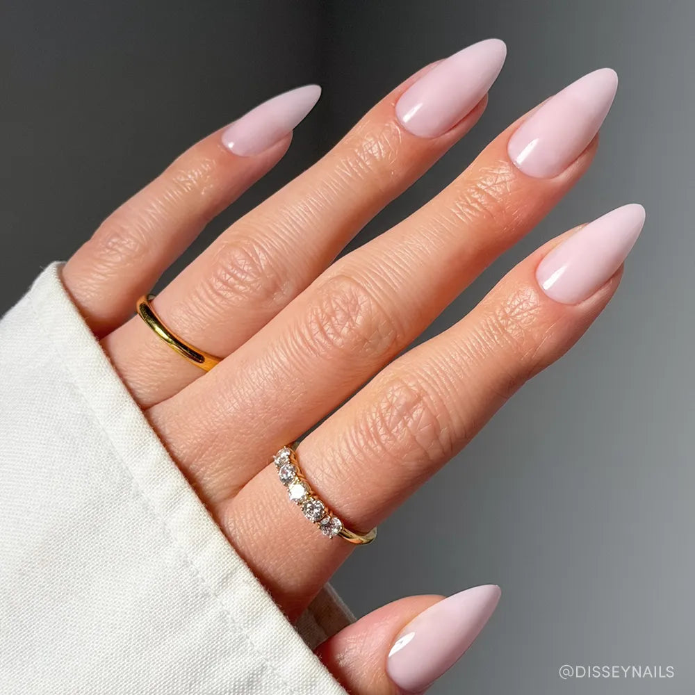 A hand with long, almond-shaped pale pink nails is shown wearing gold and diamond rings with a cream sleeve, highlighting glamnetic’s Blush Blanket Press-On Nails for an elegant, vegan, cruelty-free manicure.