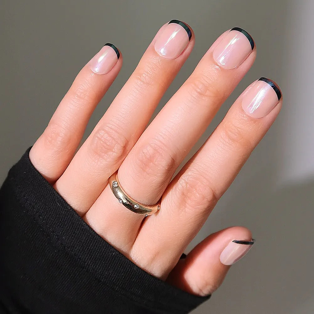 A hand with short, natural nails wears glamnetic's Burnt Miso, a minimalist French manicure with thin black tips and a glossy finish. The person sports a gold ring and a black long-sleeve top.