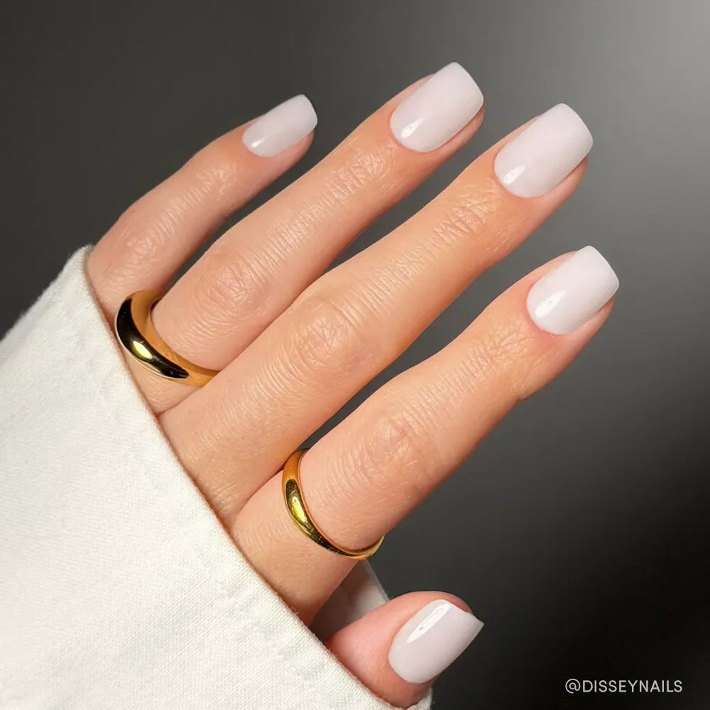A hand with light pink, glossy, square-shaped Clean Cut nails by glamnetic features a milky mani effect and is adorned with two gold rings. The hand rests on a white sleeve against a dark background.