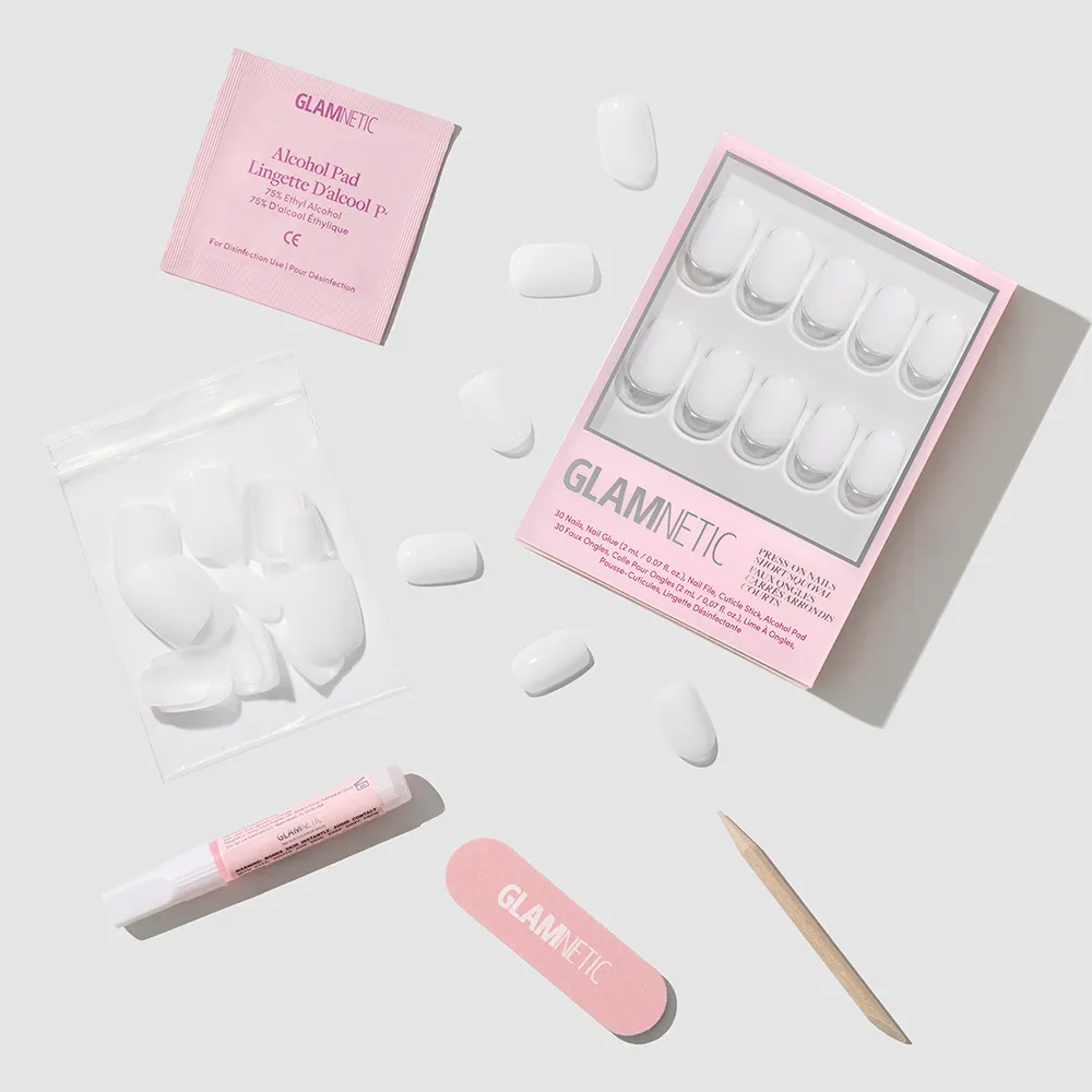A flat lay of the glamnetic Clean Cut press-on nails kit, showing a box of white milky nails, extra nails in a pouch, pink nail file, cuticle stick, nail glue, and alcohol pad on a light grey background.