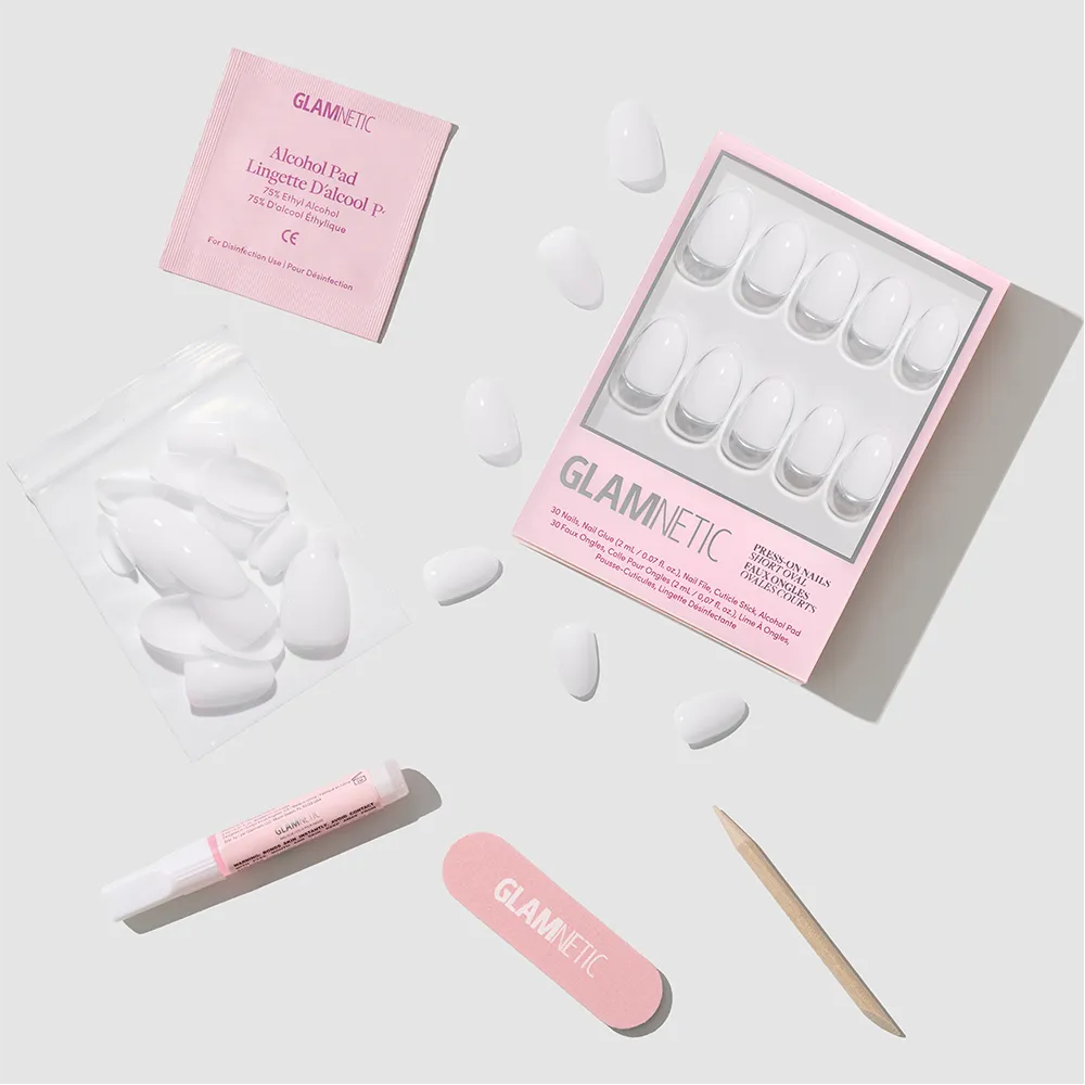 The glamnetic Clean Girl set includes milky white press-on nails, a pink nail file, a wooden cuticle stick, glue, an alcohol pad, and an extra pouch of short oval nails arranged neatly on a light gray surface.
