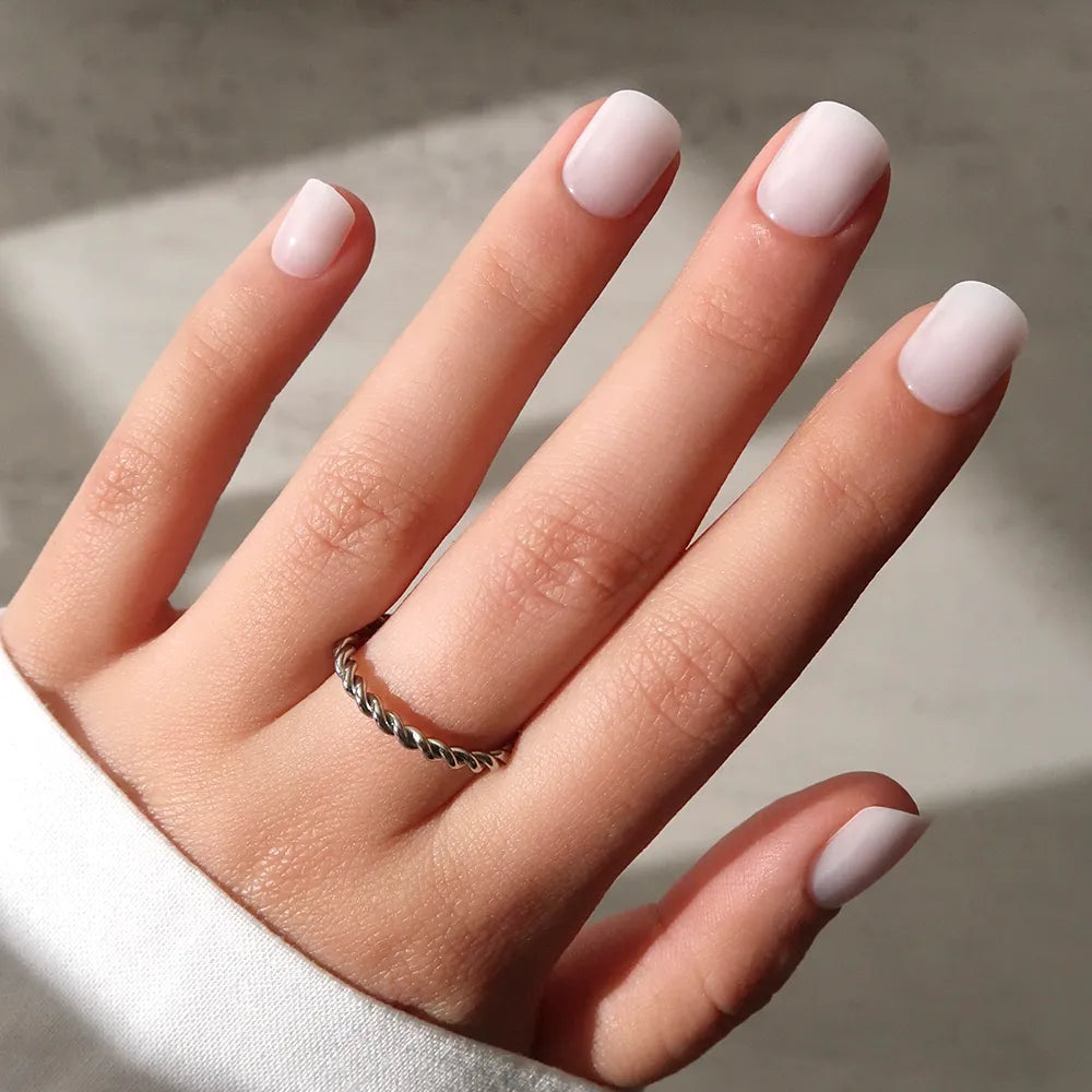 A hand with super short, square-shaped nails features the glamnetic Cool Whip mani in light pink milky color. A twisted silver ring adorns the middle finger and a white sleeve partially covers the wrist, set against a neutral background.