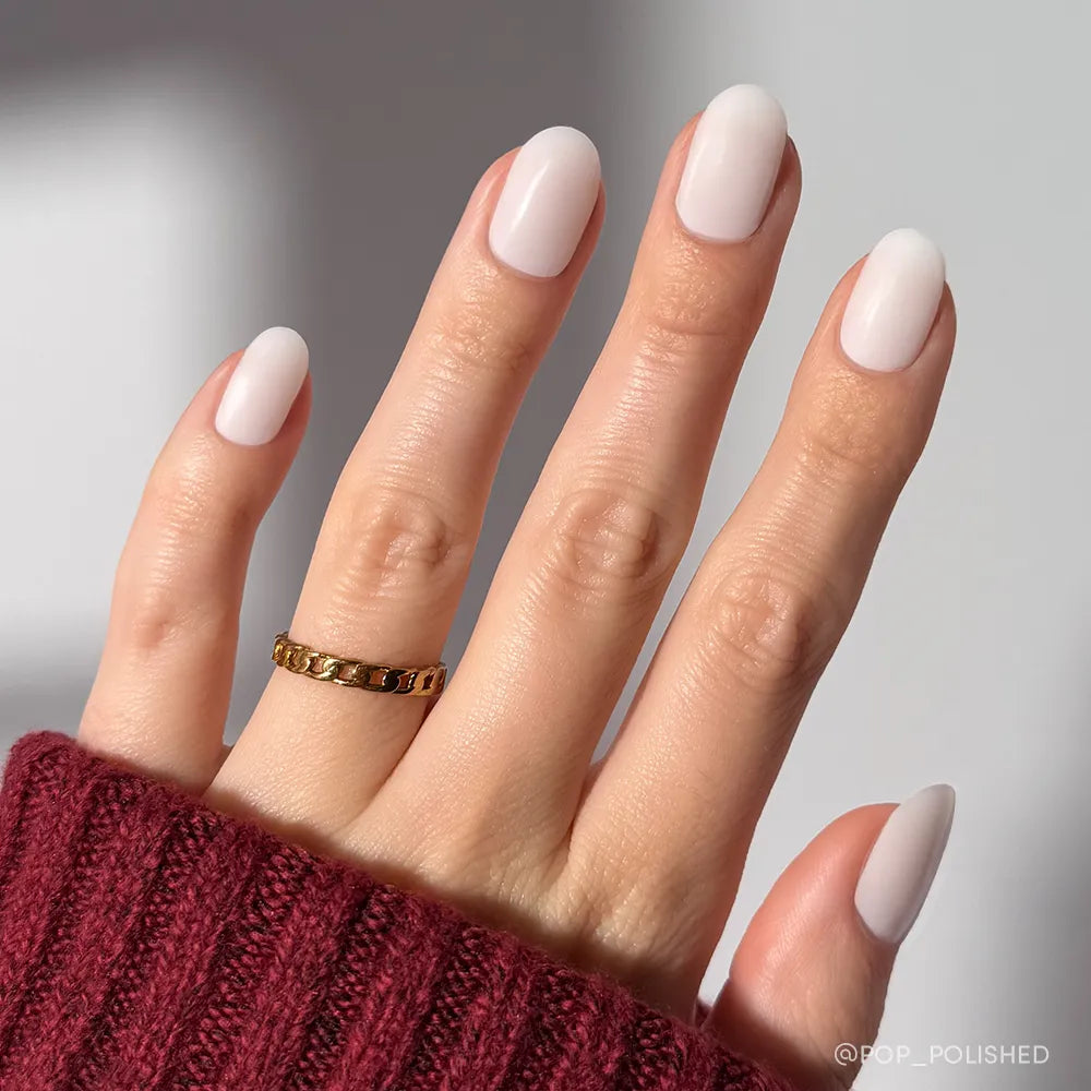 A hand with neatly manicured, short round nails features the soft, pale pink "Milk Bath" mani by glamnetic. The person wears a maroon ribbed sweater and a gold chain ring on their middle finger.