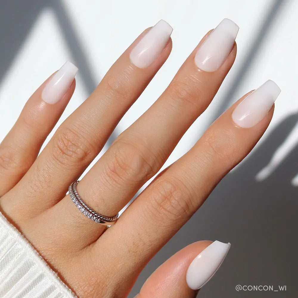 A hand with long, coffin-shaped nails features glamnetic's Oat Milk polish—a glossy, pale pink ombre for a chic milky mani. Two thin silver rings adorn the ring finger against a softly lit background with gentle shadows.