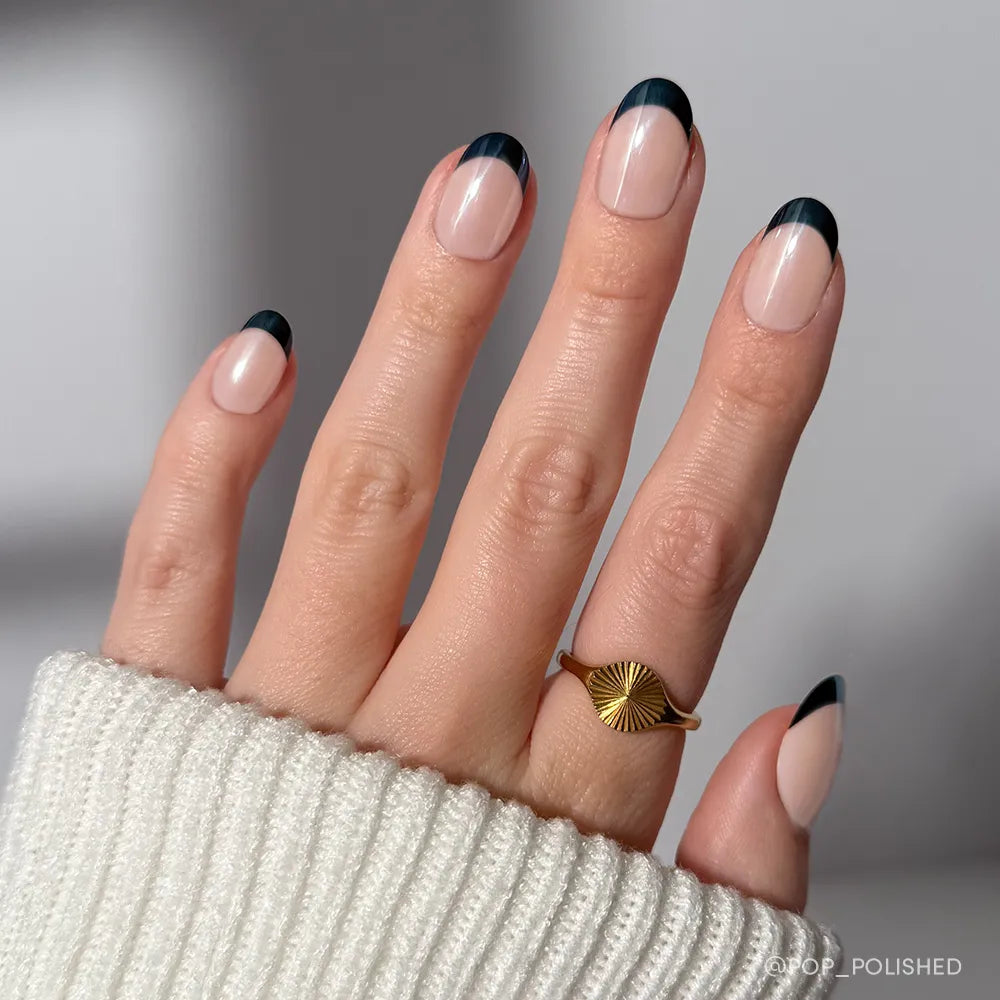 A hand showcases glamnetic's Onyx Glaze press-on nails with minimalist black French tips and a glossy finish. A gold sunburst ring and a white ribbed sweater sleeve add an elegant touch to the look.