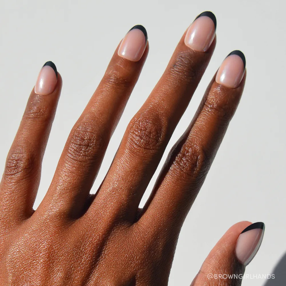 A hand with medium brown skin displays glamnetic Smoke Salt short oval press-on nails, featuring a nude base and thin black French tips, set against a white background.