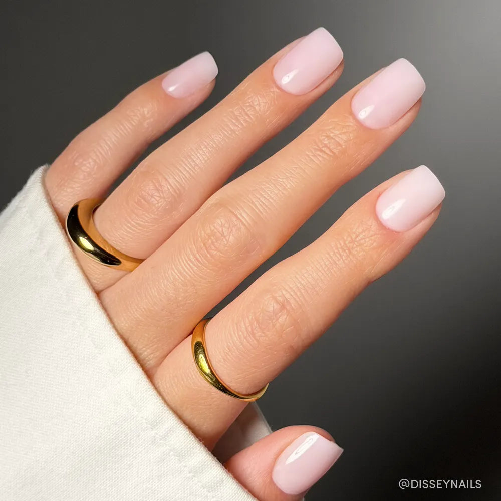 A hand with short squoval nails painted in glamnetic’s Soft Girl, a glossy pale pink. The salon-quality mani is paired with two thick gold rings and partly covered by a white sleeve.