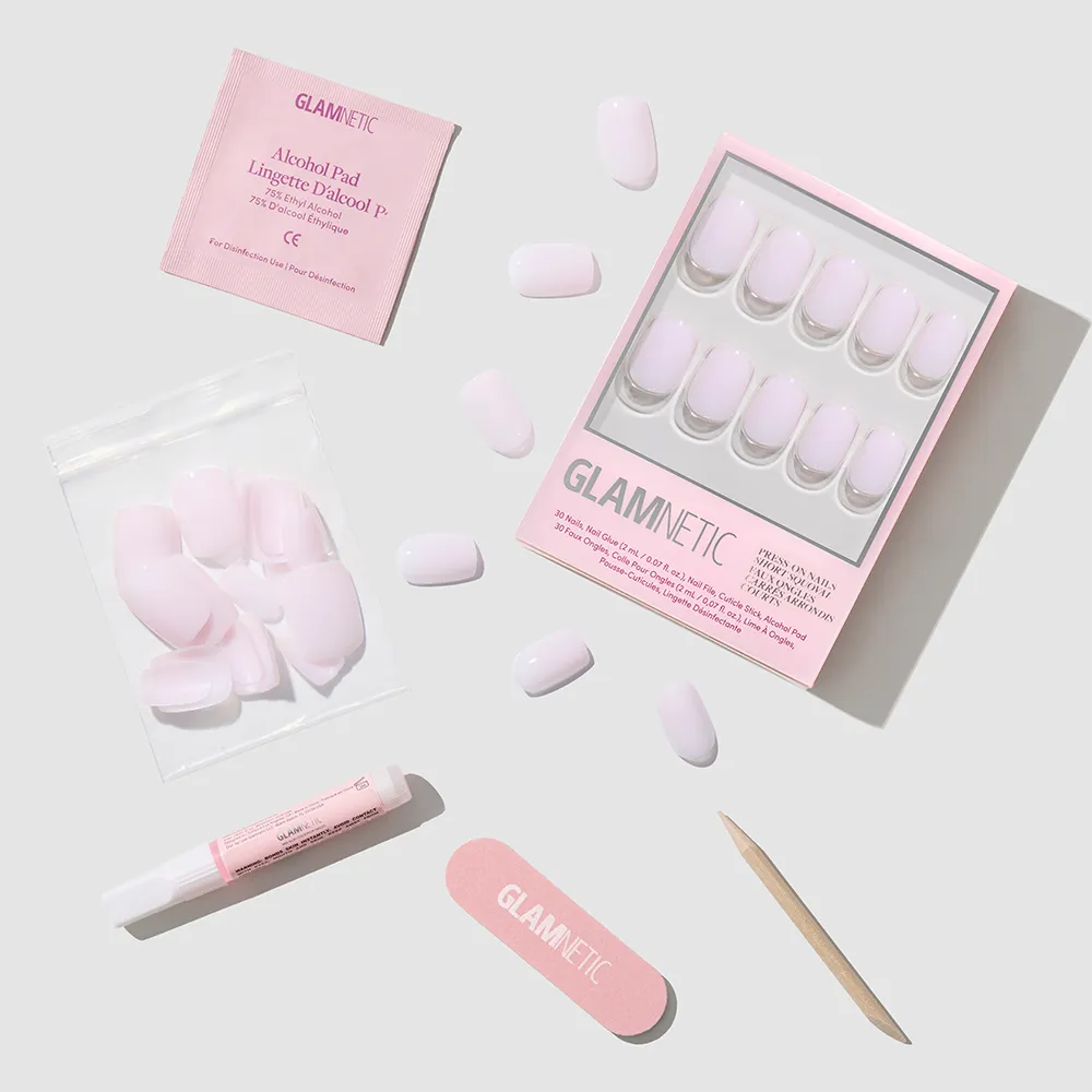 A set of glamnetic Soft Girl pink press-on nails in a short squoval shape, with nail file, cuticle stick, glue, extra nails, and alcohol pad arranged on a light gray background for a salon-quality mani at home.