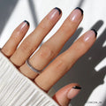 A hand with a light skin tone features glamnetic’s Squid Ink minimalist black French tip nails with a glossy finish, accessorized by two thin sparkly silver rings and a white pleated sleeve.