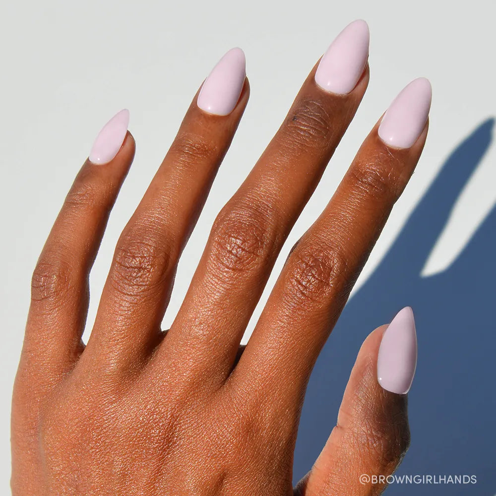 A hand with medium brown skin shows short almond nails painted glossy pink using glamnetic’s Sugar Doll press-ons, set against a pale background.
