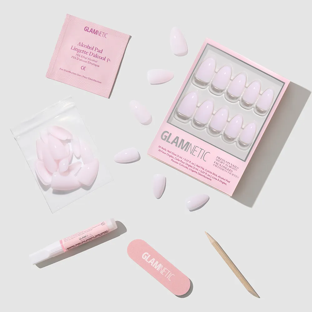 The glamnetic Sugar Doll set features glossy pink short almond press-on nails and includes nail glue, an alcohol pad, a wooden cuticle stick, a nail file, and extra nails—all arranged neatly on a light gray background.