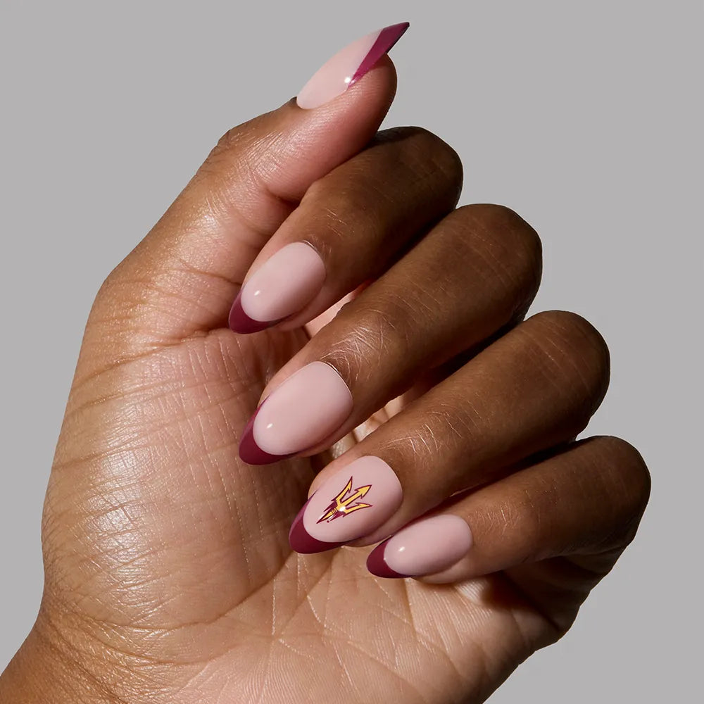 A hand with almond-shaped glamnetic Arizona State Sun Devils Press-On Nails featuring a nude pink base, maroon French tips, and a gold lightning bolt on the ring finger, shown against a plain gray background.