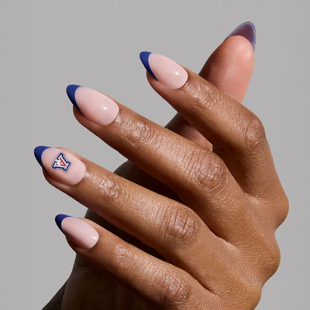 A hand showcases almond-shaped, salon-quality nails with a nude base and navy French tips from the glamnetic Arizona Wildcats Press-On Nail Set; the thumb features a red, blue, and white Wildcats V logo against a light gray background.