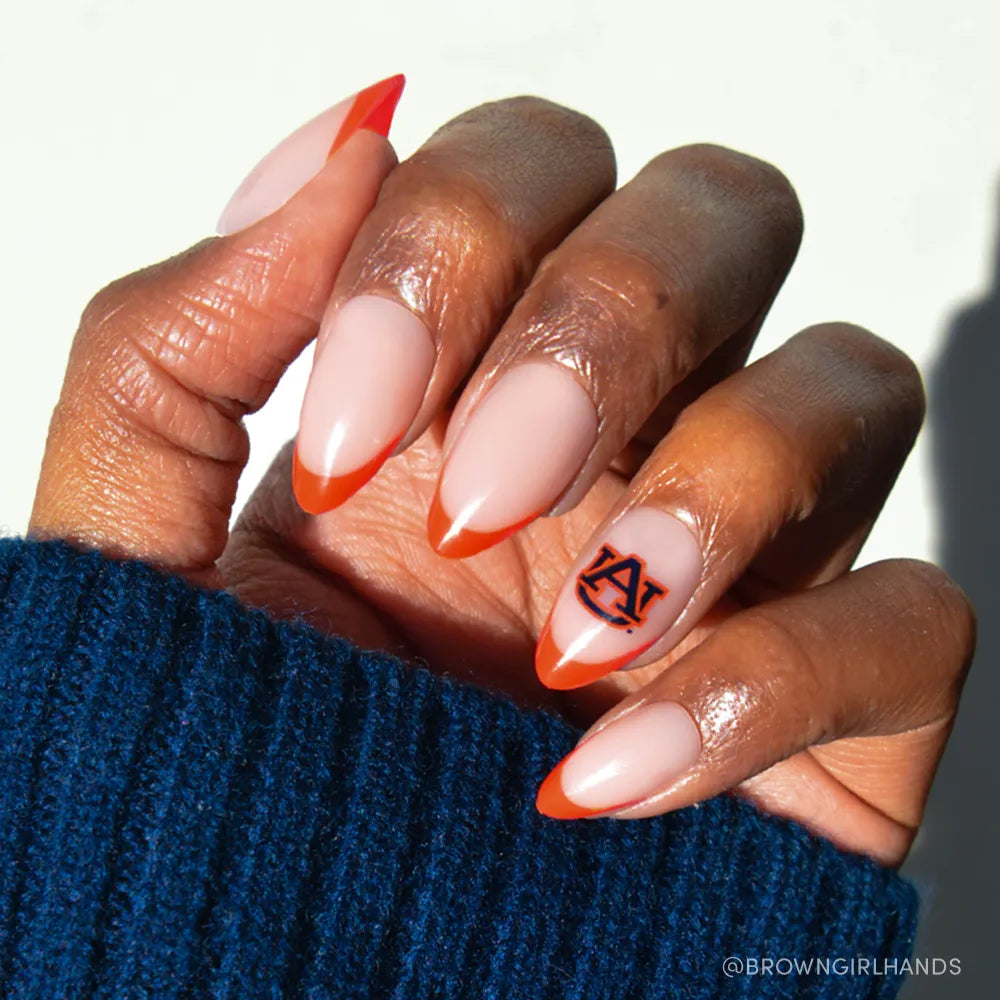 A hand wears glamnetic's Auburn Tigers Press-On Nail Set, with almond-shaped nude nails and bright orange French tips. The ring finger features the team's blue and orange logo. A blue knit sleeve is worn.